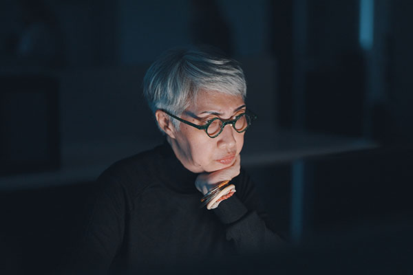 older woman of Asian descent sitting in a darkened room lit by the light of her laptop